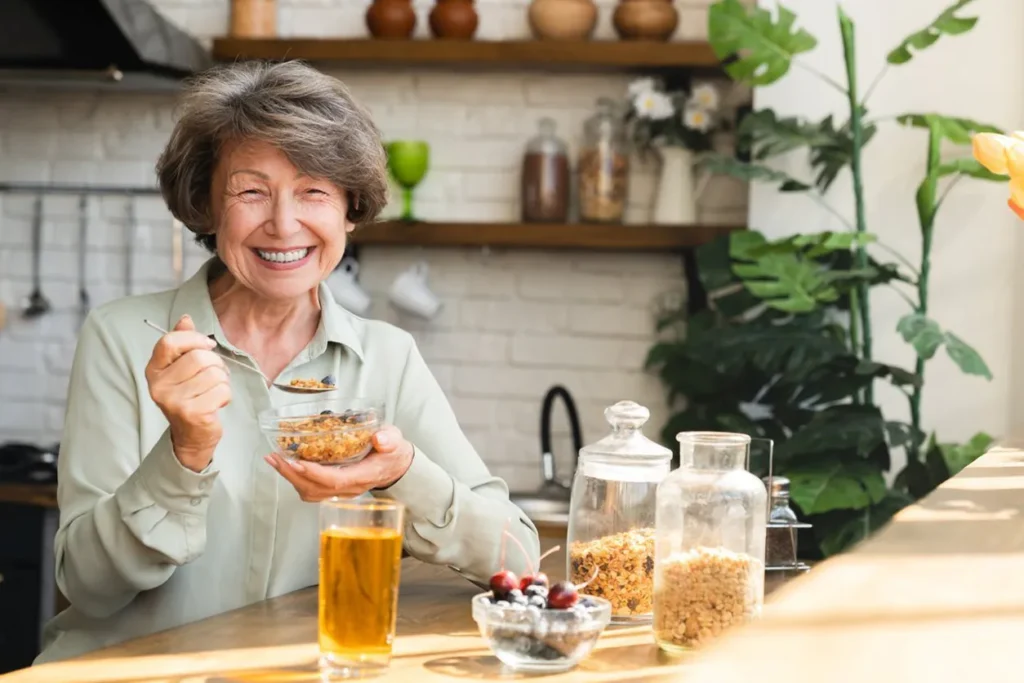 A smiling senior woman eats a bowl of oats in the kitchen.