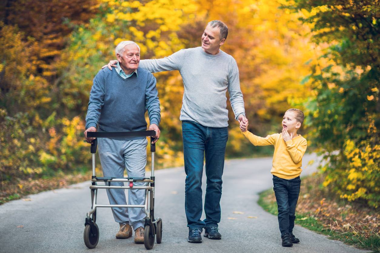 A senior father with a walker, an adult son, and a young grandson walk together in the park