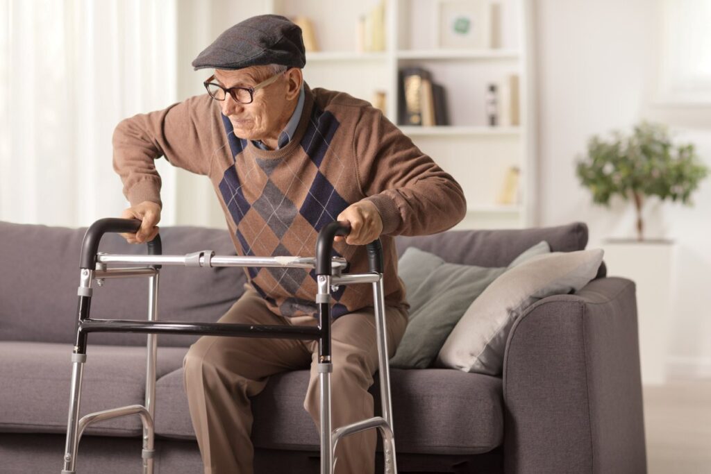 An older gentleman in an argyle sweater and a hat uses a walker to get off the couch.