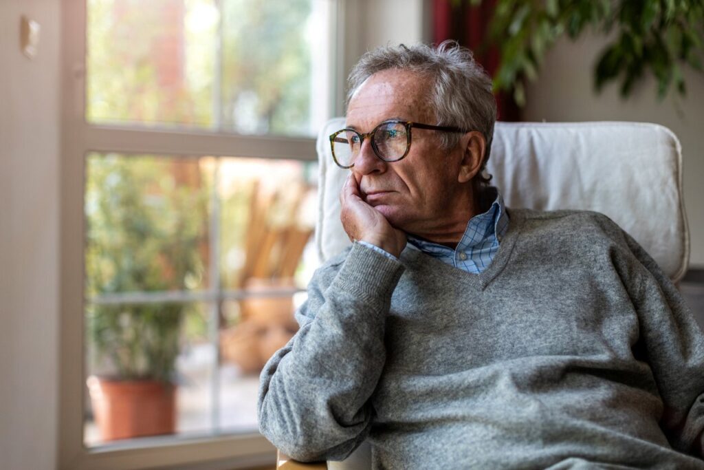 An older man with his hand under his chin stares out of the window at home.
