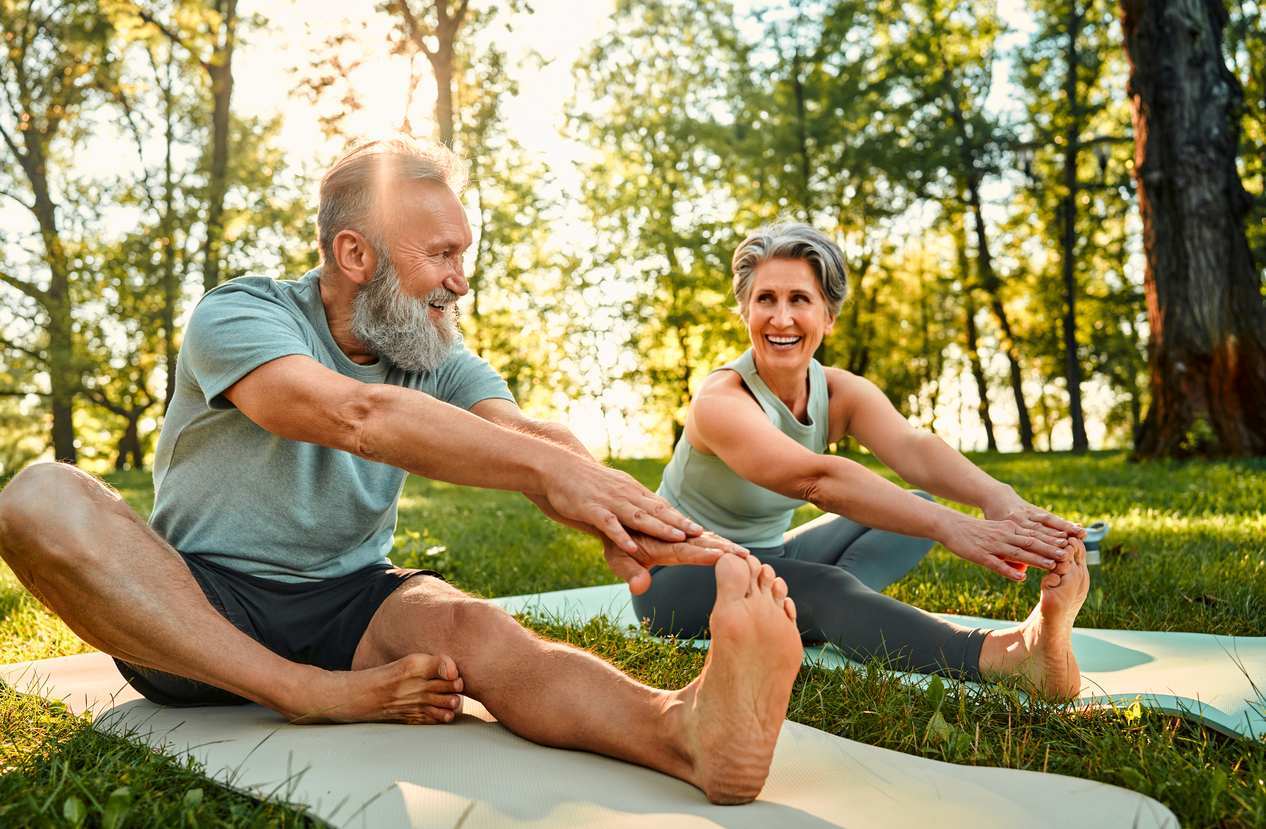 Two seniors stretch on yoga mats in the woods on a sunny day.