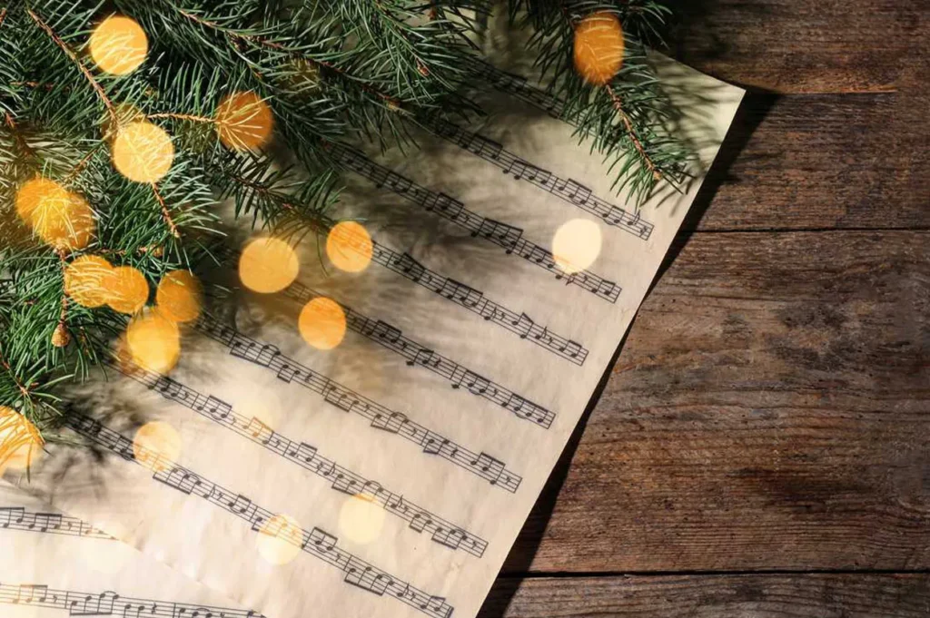A sheet of Christmas music placed on a wooden surface with garland and lights surrounding it.