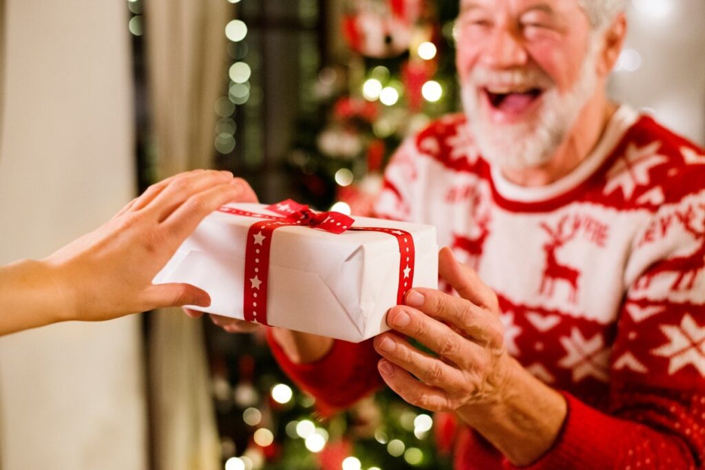 An older man receives a Christmas gift while wearing a festive red and white sweater in a decorated living room.